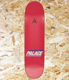 A red Palace skateboard deck shown vertically against a background of shredded wood chips. The deck features the Palace logo in white and blue near the bottom, and a black triangle logo near the top.